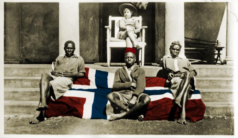 Kolorert bilde. Et norsk flagg er brettet ut over en trapp til en balkong, tre afrikanere sitter på flagget. En lys gutt sitter på en stol på balkongen.