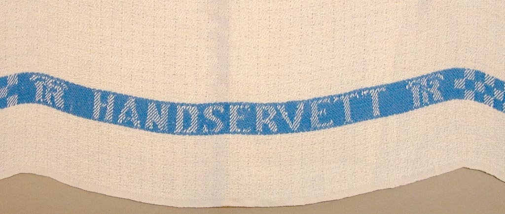Handduk eller handservett i vitt linne med en vävd blå bård med texten "TR handservett TR" invävt i tyget.