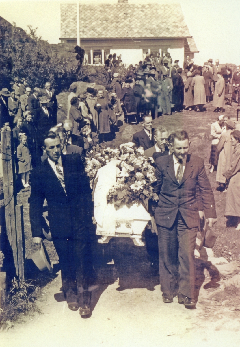 Begravelse. Seks menn bærer en hvit kiste med blomsterkranser på. Mange sørgende står langs ruten. Hvit våningshus på høyden i midten bak. Hekk til venstre.