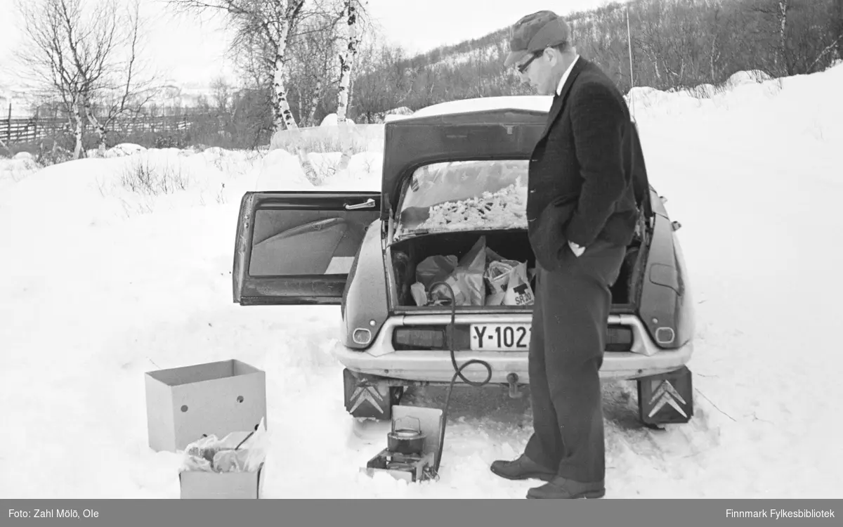 En Citroën bil parkert lang veien. En mann koker kaffe med "Primus" (tur/frilufts-kokeapparat). Hvor er bildet tatt, kanskje et sted i Tana? Tidspunkt er rundt 1970.