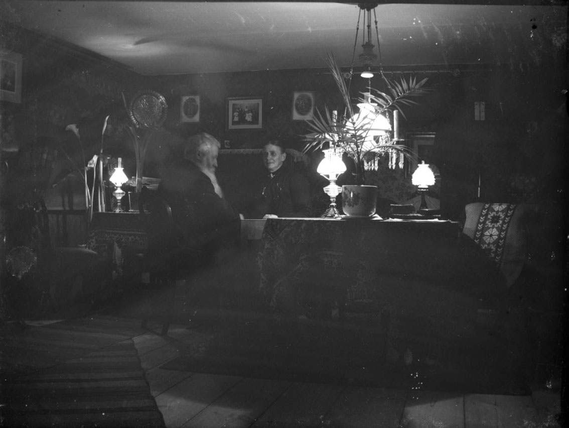 Interiör från ett okänt hem med ett okänt äldre par. De sitter vid ett bord, det är kväll. Flera fotogenlampor är tända, i tak och på bord.