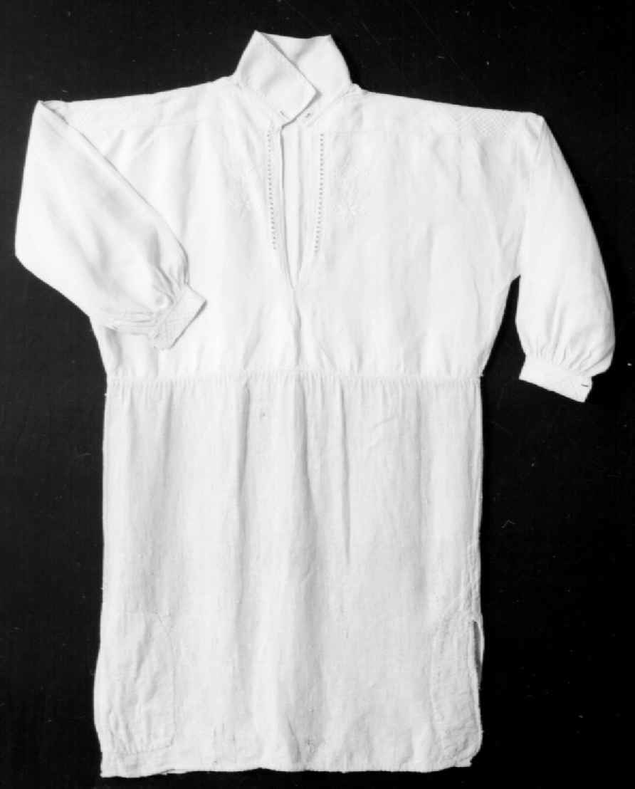 Bunadsskjorte av lin, med brodering og hullsøm på mansjetter, krage og stolpe. Nederste del av skjorten er av strie.