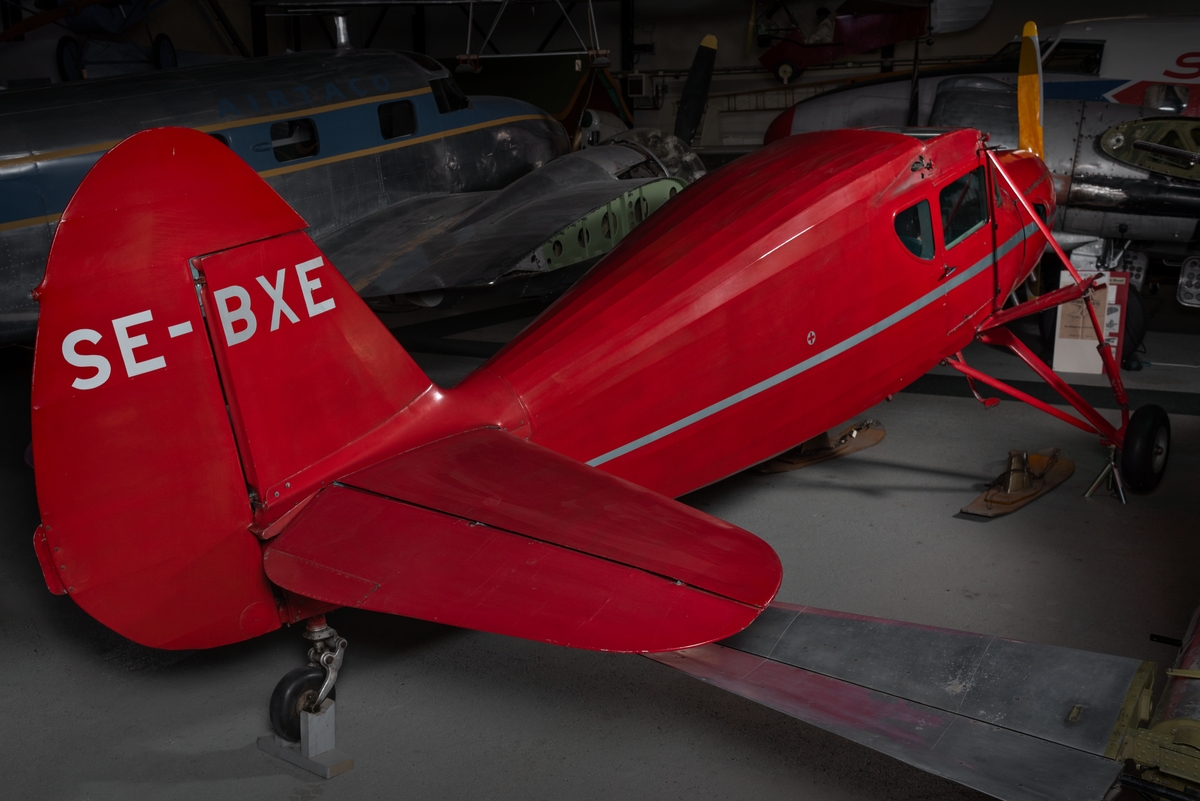 Flygplan av modellen Fairchild 24. Rödmålat propellerflygplan med silverfärgad pil på flygkroppens båda sidor. Propeller av trä.