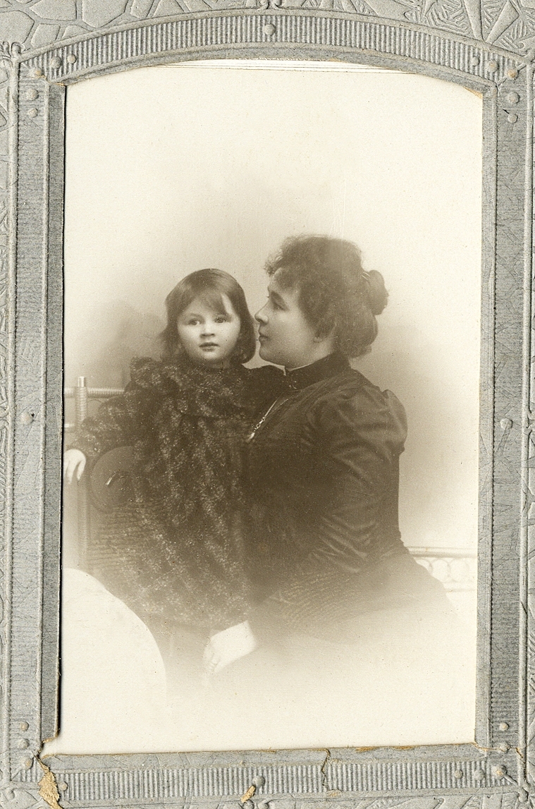 En kvinna i mörk klänning med hög krage och en liten flicka i mörk, rutig klänning. 
Knäbild. Ateljéfoto.

Fotografen med dotter.