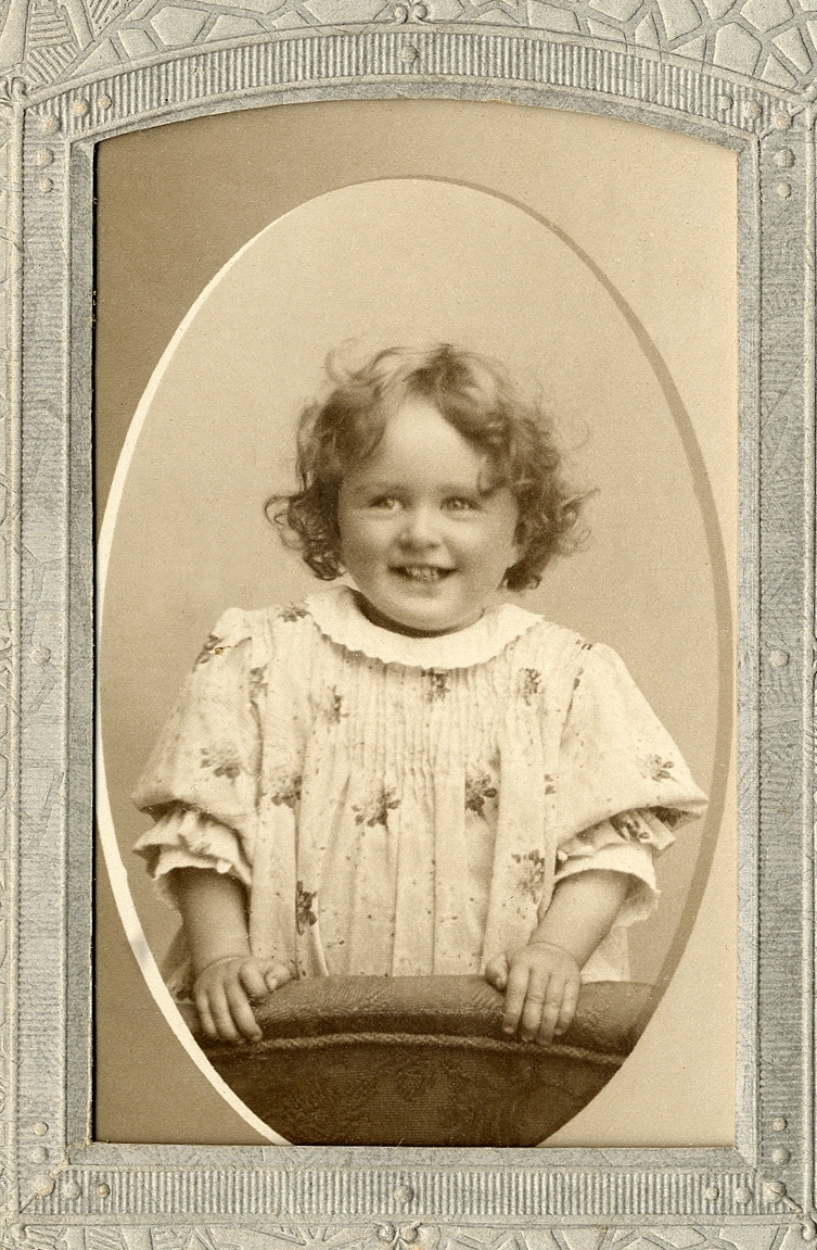 En liten glad flicka i småblommig klänning, som vilar händerna mot ryggstödet på en stoppad stol. 
Knäbild. Ateljéfoto.

Fotografens dotter.