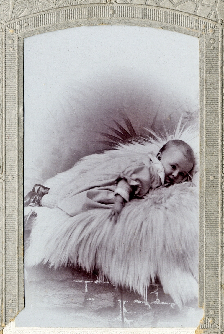 En baby i kolt som ligger på en raggig fäll. 
Helfigur. Ateljéfoto.

Fotografens dotter.