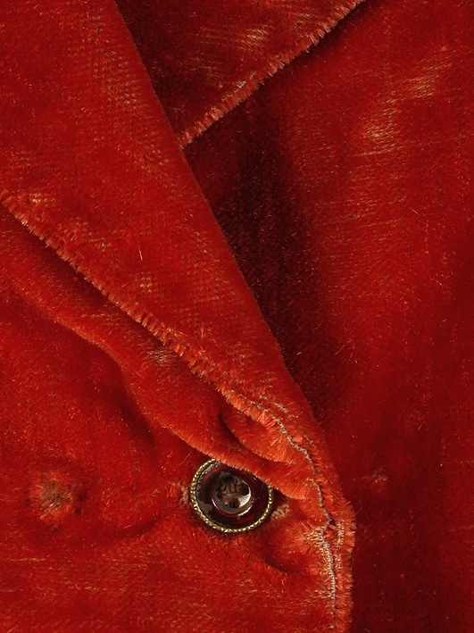 Röd sammetsplysch, krage och slag. Två stolpfickor. Rygg av beige linne. Dubbelknäppt med fyra knapphål på vardera framstycket.
