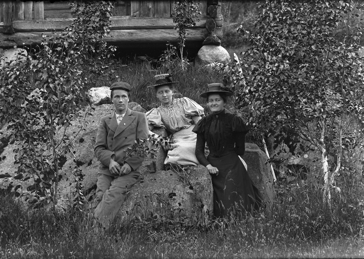 En mann og to kvinner, sitter ute på en stor stein. Bjørker rundt.