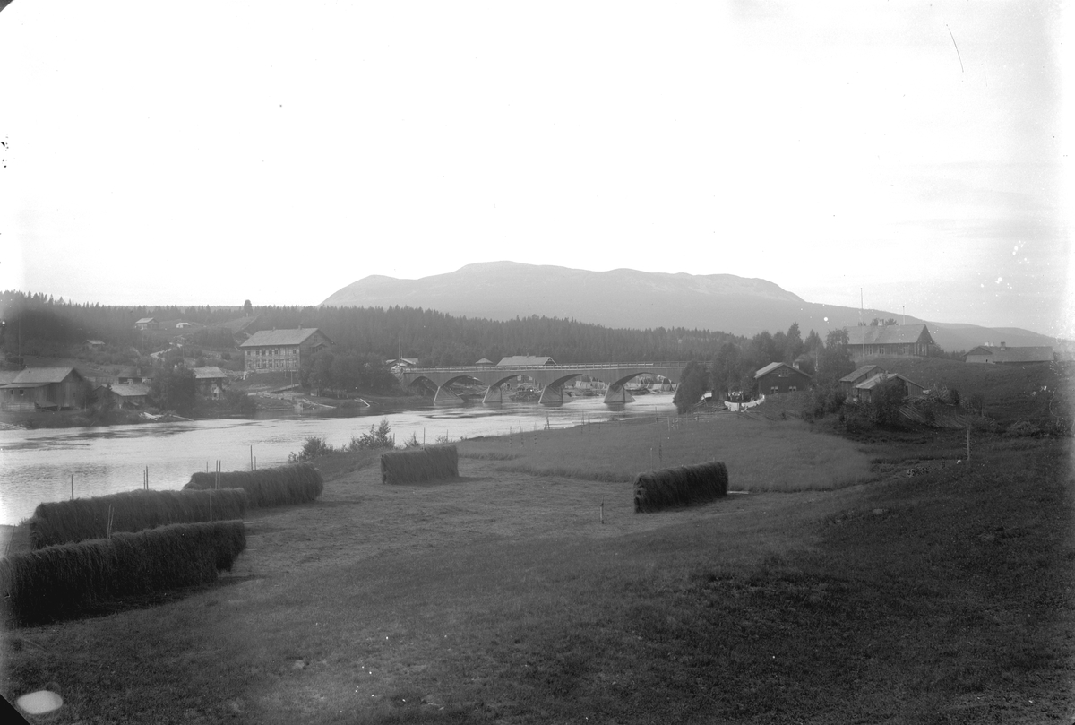 Nybergsund. Fotografert fra østsida av Trysilelva mot nordvest, Trysilfjellet i bakgrunnen. Brua sto fra 1879-1929. På vestsida av elva ses Nybakk og Nyberg, huset i to etasjer på østsida som ligger delvis skjult bak en høyde er Folkvang.