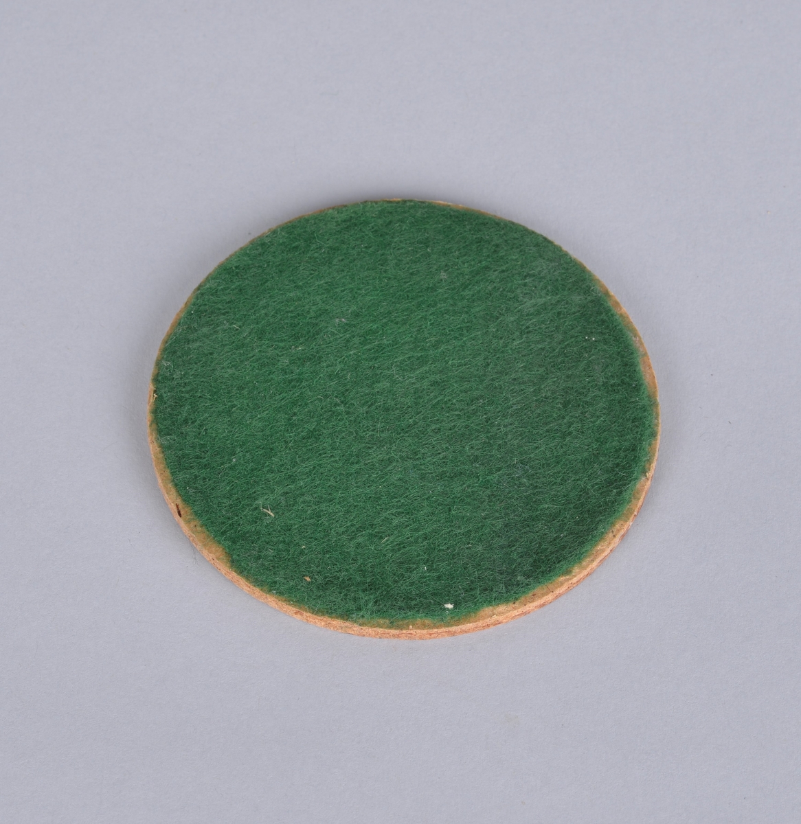 Rund trefiberplate (huntonitt?) med pålimt grønt filtbelegg.
Ukjent bruksområde.

Muligens vært brukt som underlag for glass eller utstyr på laboratoriet.