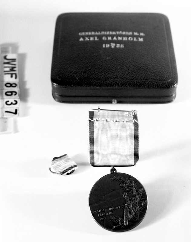 Skidfrämjandets medalj av järn, med sidenband i vitt med gul och blå kant.
Text: "Manlig idrott, fädrens arf".
