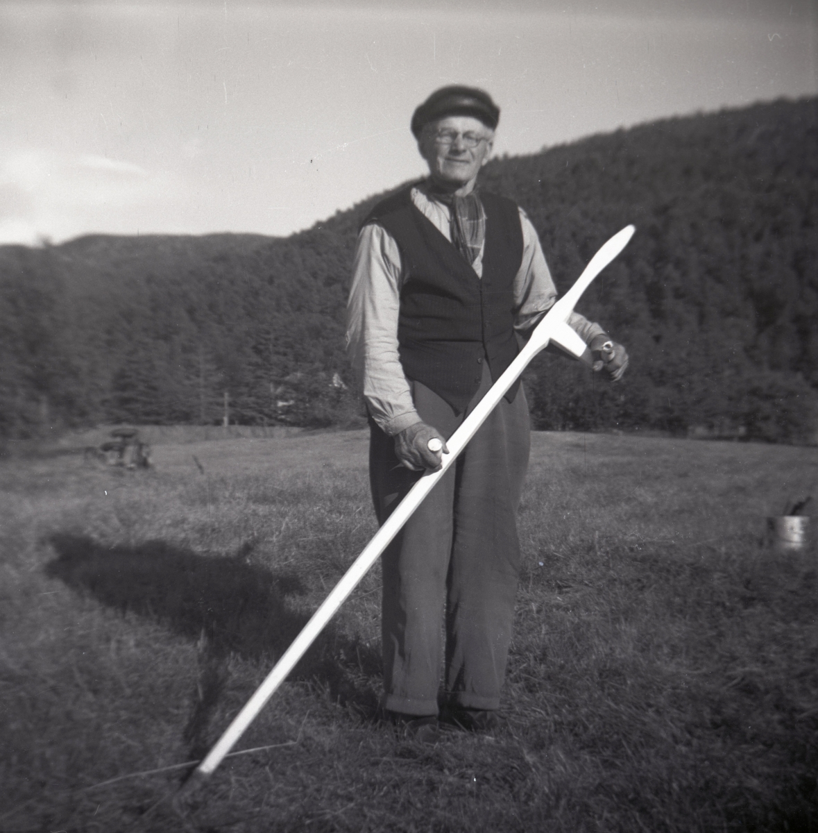 Portrettfotografi av en eldre mann med ljå som står på ei mark der det drives slåttonn.