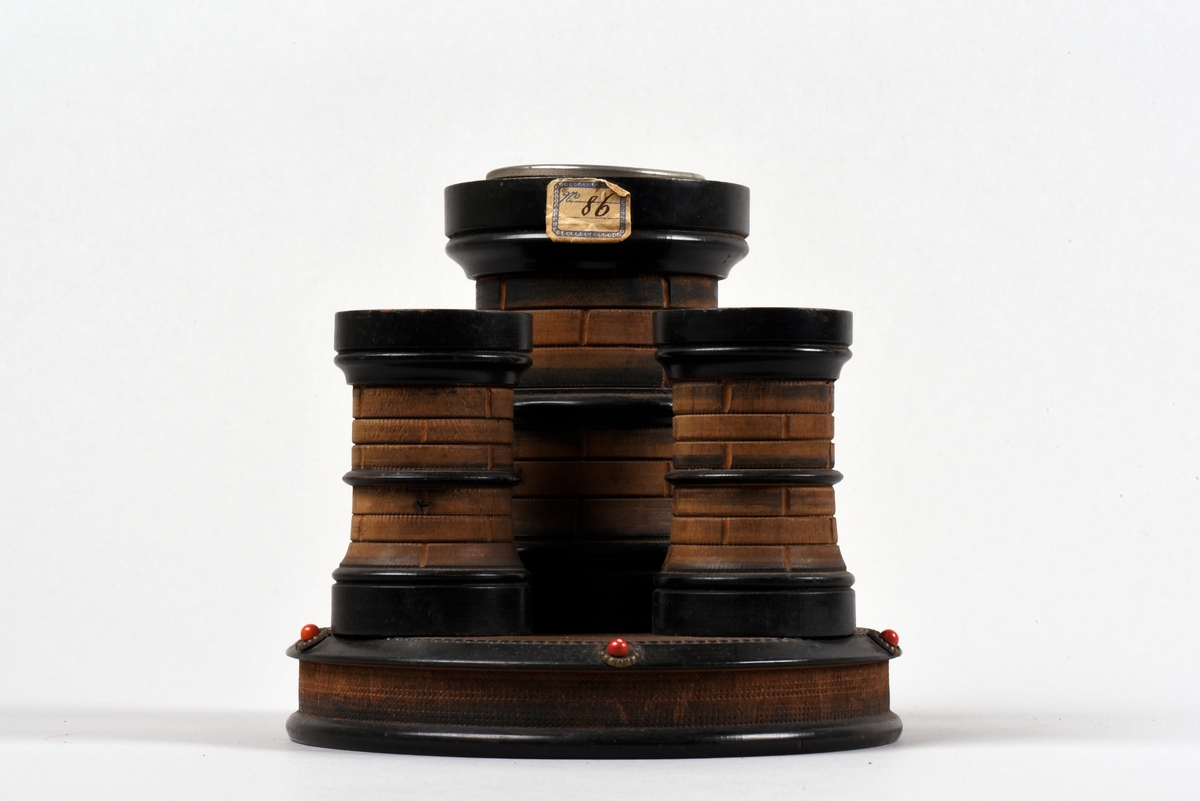 Cigarrställ av trä, i form av tre runda torn som står på en rund platta. Det högsta tornet har en metallskoning vid mynningen, de lägre olika utformade hål.