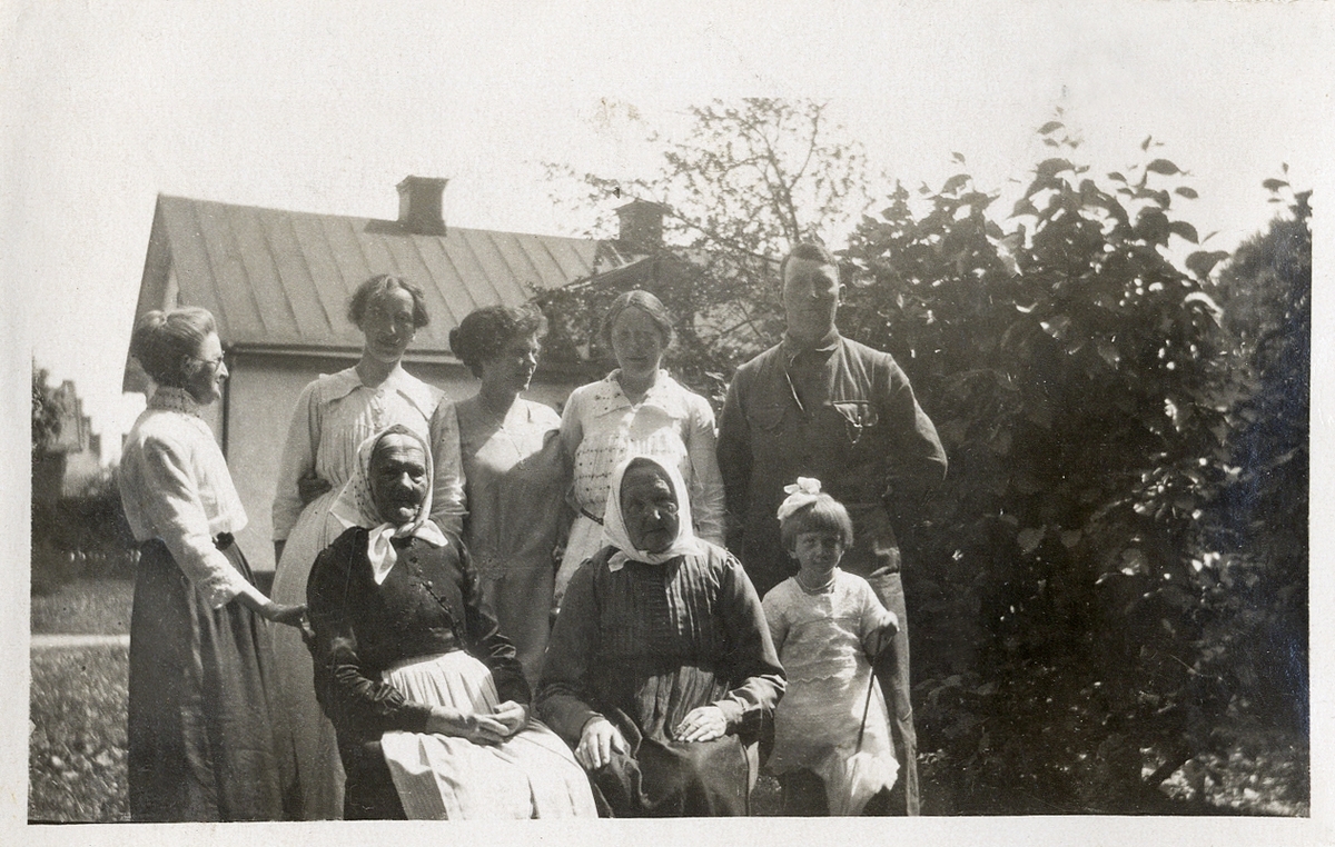 Foto av en familj med flera generationer, mestadels kvinnor. Man har samlats utomhus, i en trädgård. I bakgrunden skymtar ett bostadshus. Längst till vänster står fröken Maria Lindblom (1850-1930), Växjö.