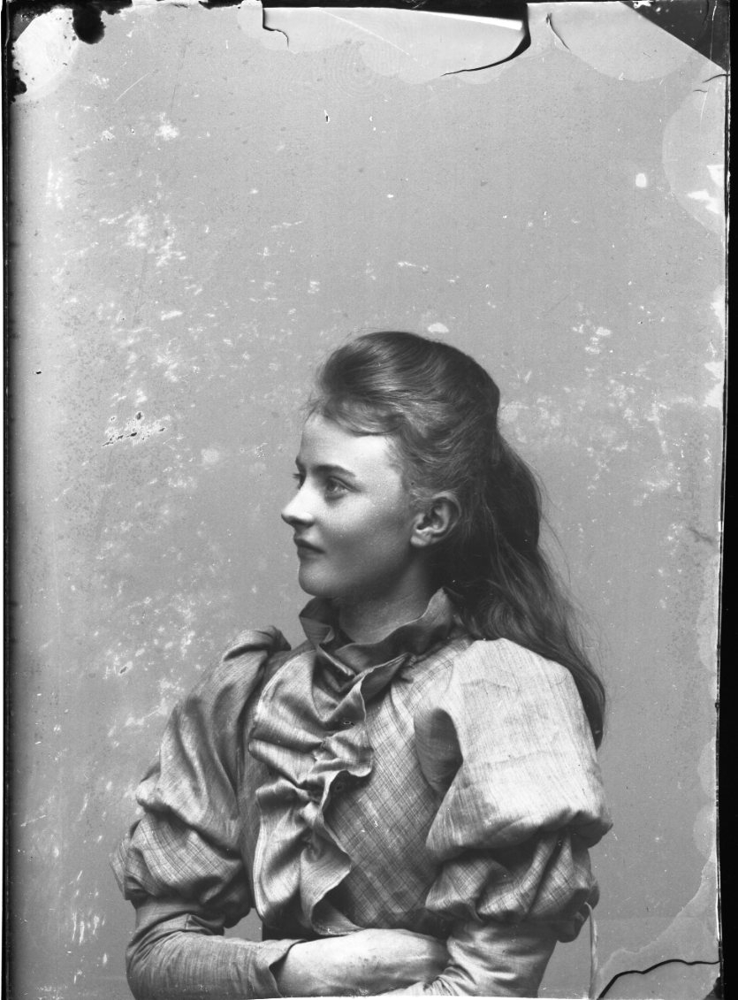 Bröstbild i 3/4-profil av Ester Grönberg. Hon bär en svagt rutig blus med krås. Hon har armarna i kors.