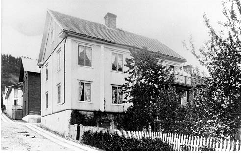 Putsat bostadshus i två våningar med ljusa lisener, i hörnet Hamnvägen-Sjögatan. Övre partiet av gavlarna har locklistpanel. Fotograferat från Hamvägen och uppåt. På balkongen till höger står tre män och tre kvinnor.