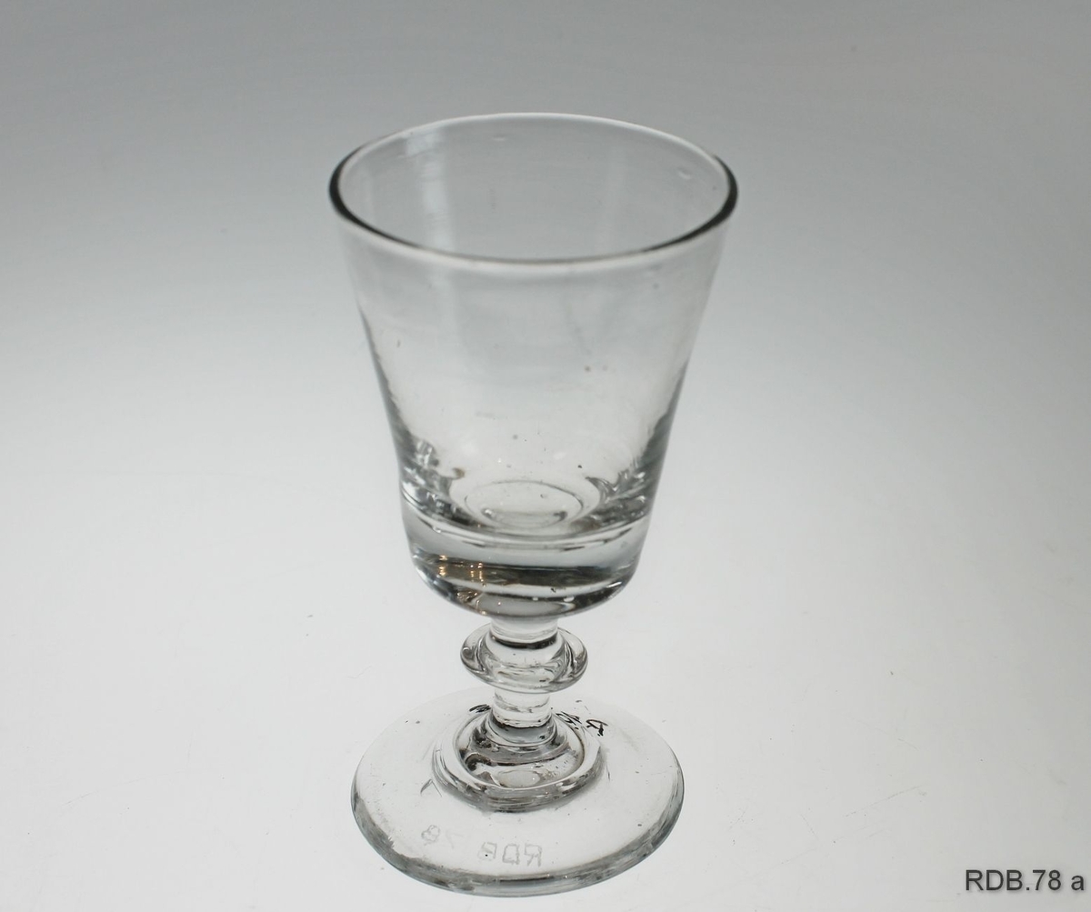 Drammeglass med "knapp" på stetten. Glasset er videre øverst enn nederst. Et merke under bunnen viser at glasset er handblåst.