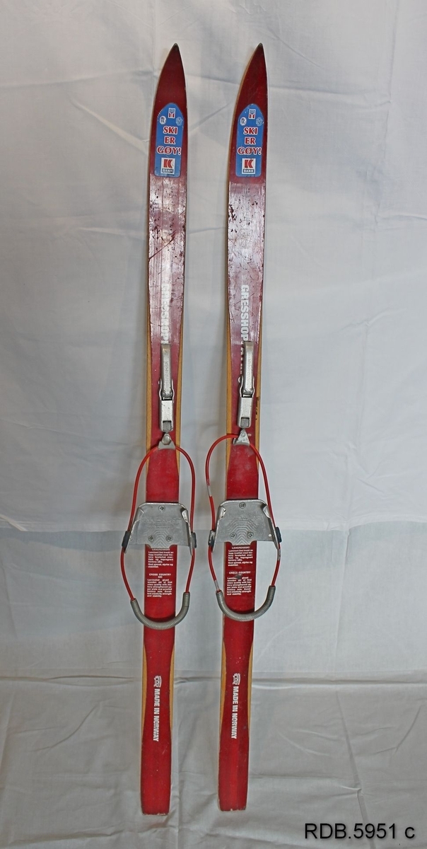 Røde barneski med med trekvite kanter. Påklistret merke foran på skia med påskrift "Ski er gøy". Kandaharbindinger. 1 igle under.