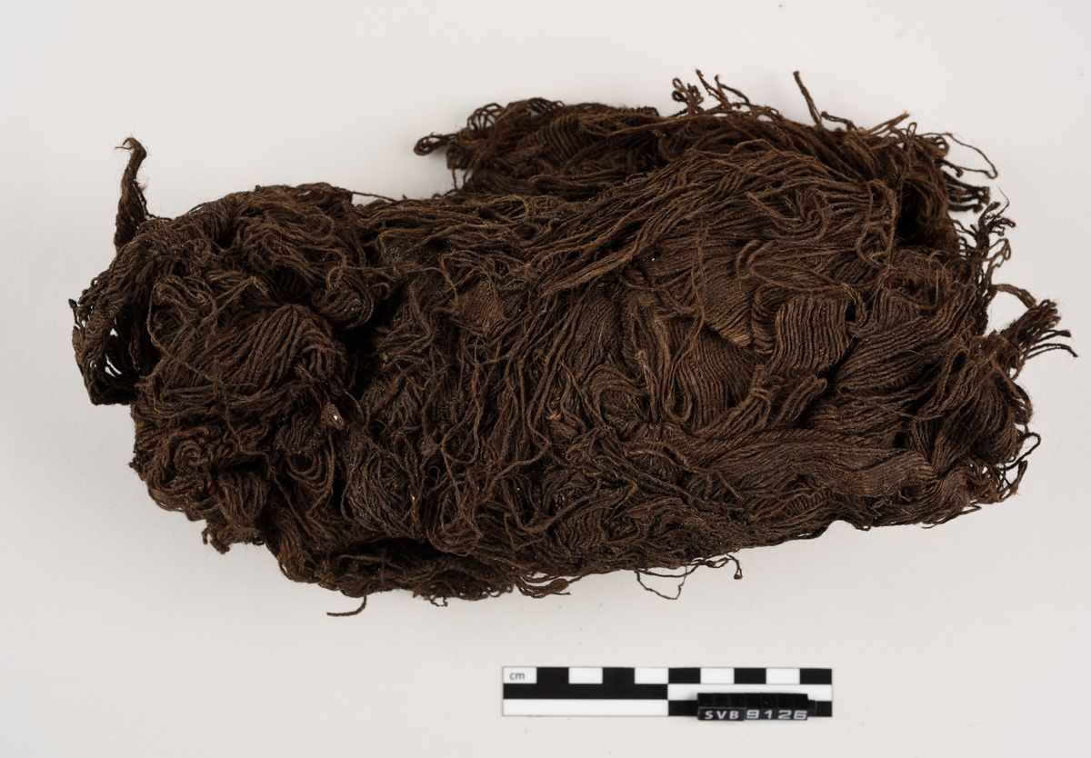 En bunt med tråd der fibrene er spunnet sammen. Det går en trepinne inn i gjenstanden. Gjenstanden er mørkebrun i farge. Gjenstanden er skjør.