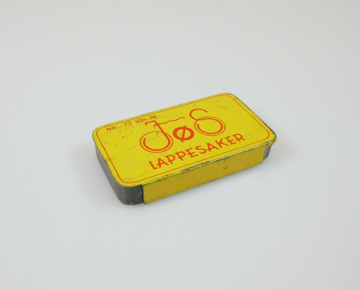 Rektangulær boks i metall. Et gult lokk som kan tas av. Boksen inneholder to deler av et svart gummimateriale og en tom limtube. 