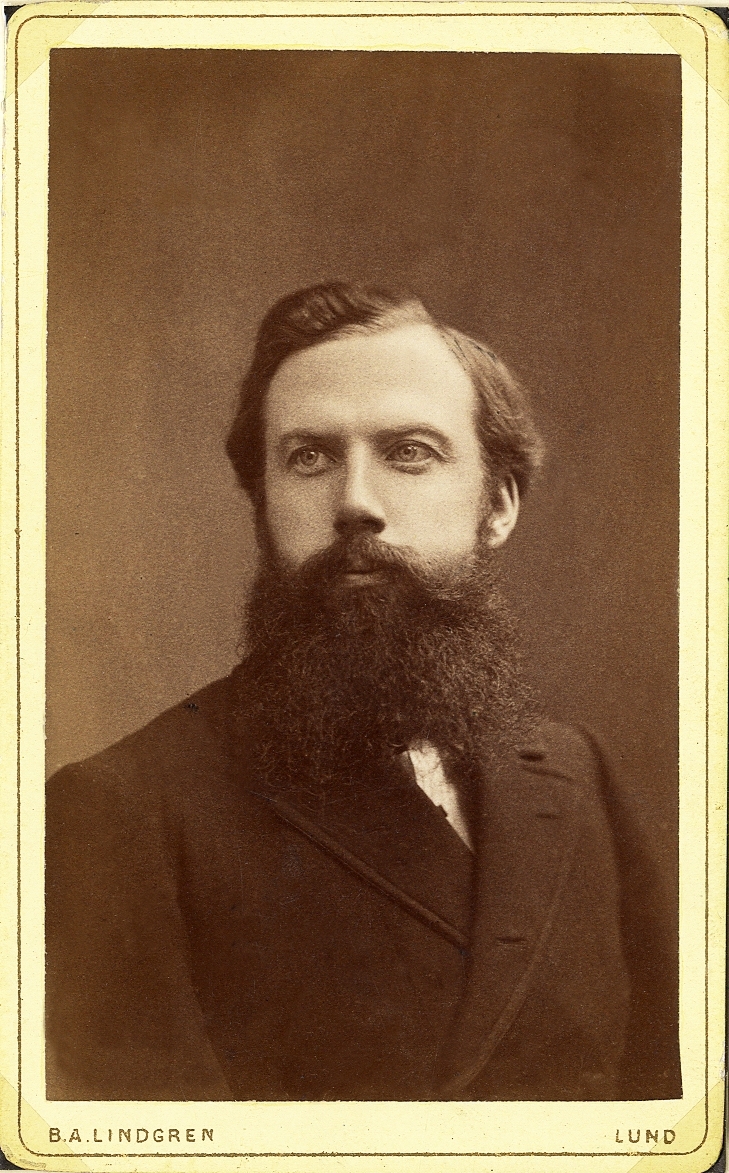 Porträttfoto av en man i helskägg, klädd i mörk bonjour (?) med stärkkrage m.m.
Bröstbild, halvprofil. Ateljéfoto.