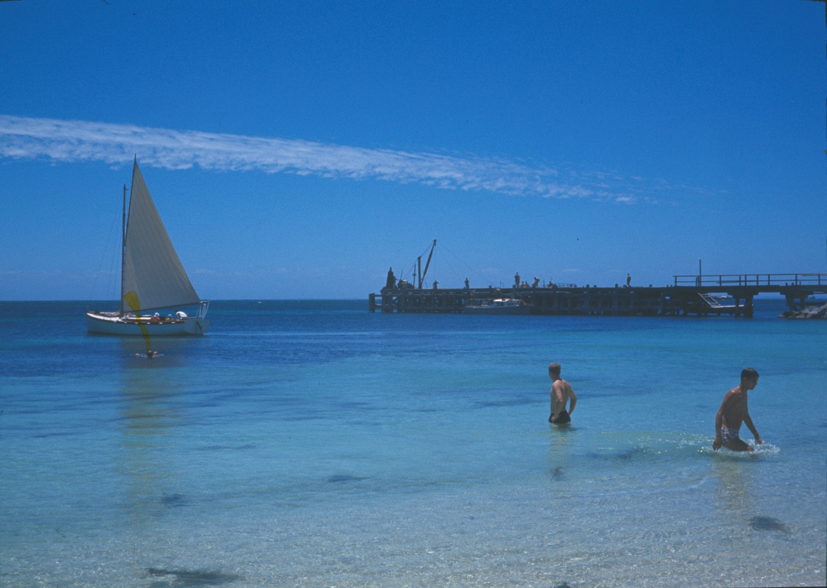 Bilden visar badgäster vid en strand i Freemantle. I viken ligger en segelbåt och i bakgrunden syns en brygga.
