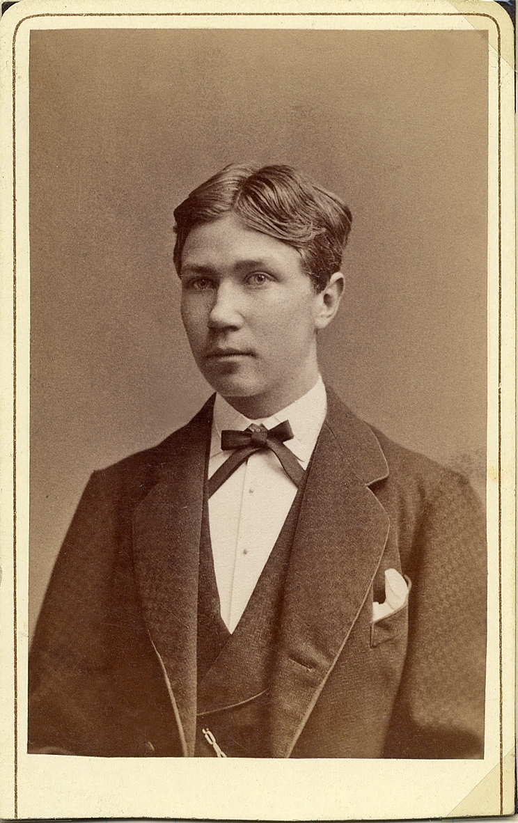 Porträttfoto av en ung man i kavajkostym med väst, stärkkrage och fluga. 
Midjebild, halvprofil. Ateljéfoto.