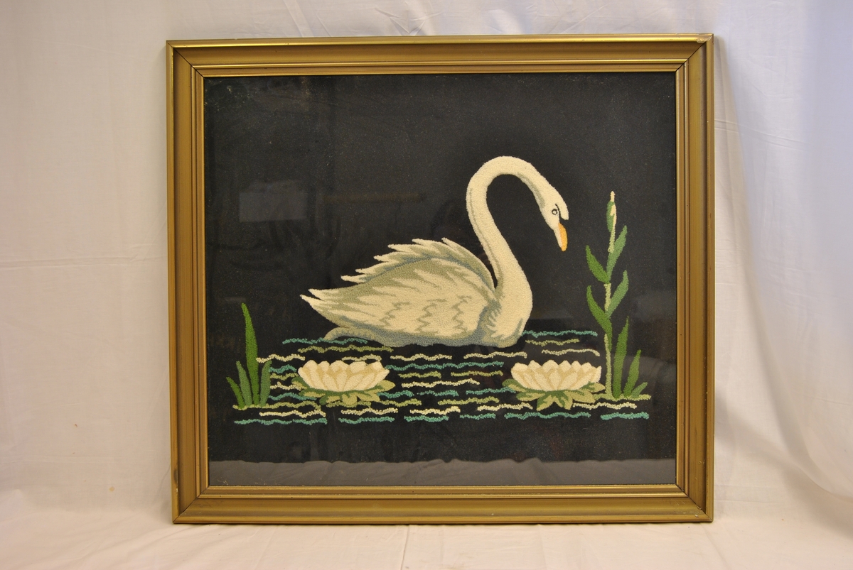 Brodert bilde i glass og ramme. Rammen er forgylt. Bildet forestiller en svømmende svane mot sort bakgrunn. 