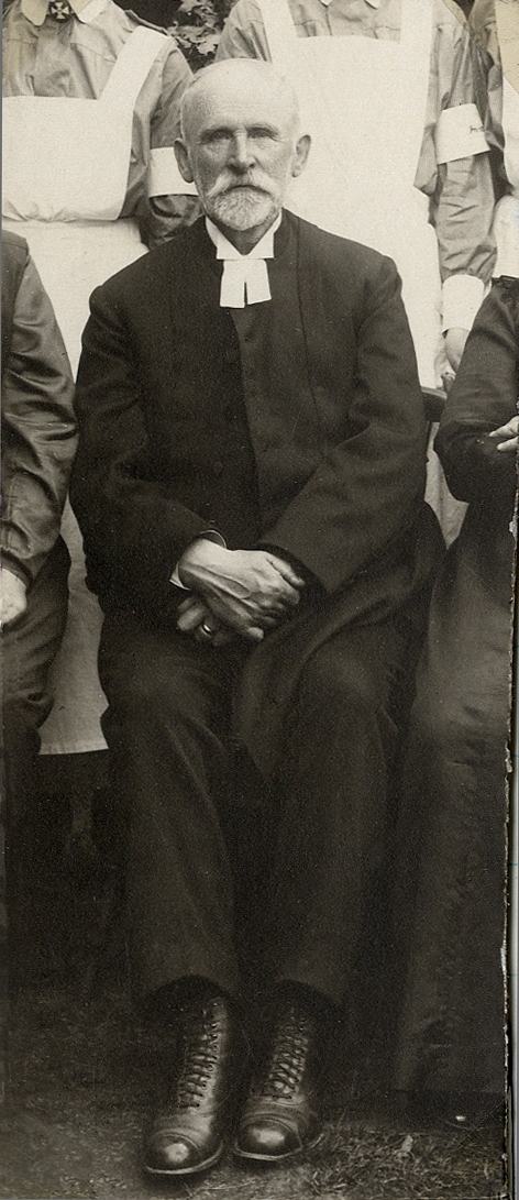 Foto av en äldre man med mustascher och pipskägg, klädd i prästrock och prästkrage.
Utsnitt ur större foto.