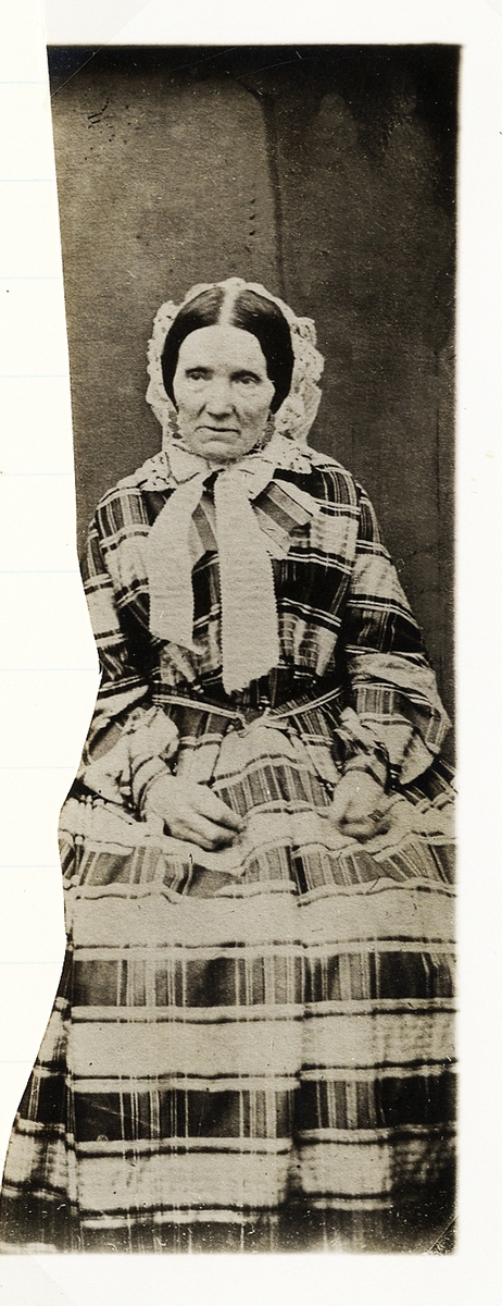 Foto av en äldre kvinna i tvåfärgad bredrandig krinolin med spetsmössa på huvudet. 
Helfigur, en face. Ateljéfoto.