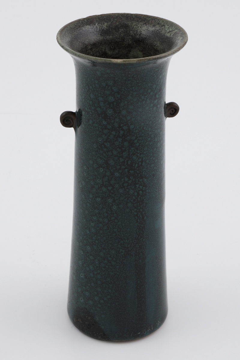 Avlang sylinderformet vase av steingods. To små volutter er plassert på hver side av øvre del av korpus. Yttersiden er dekket av en blåfarget glasur spettet med sort, mens innersiden er grågrønn.  Stemplet: "Auguste Delaherche / 1897".