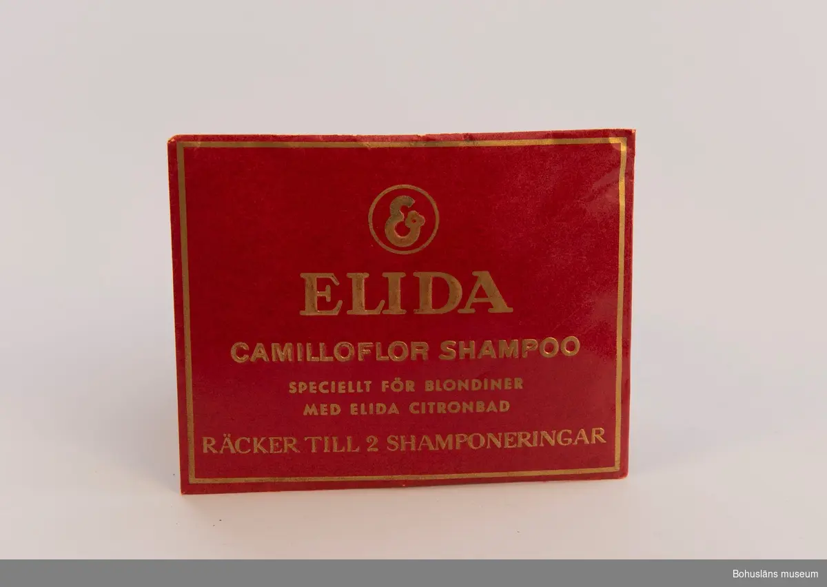 Papperskuvert med pulvershampoo. Text i guldfärg mot vinröd botten: "Elida. Camilloflor shampoo. Speciellt för blondiner med Elida citronblad. Räcker till 2 shamponeringar." 
På baksidan bruksanvisning.