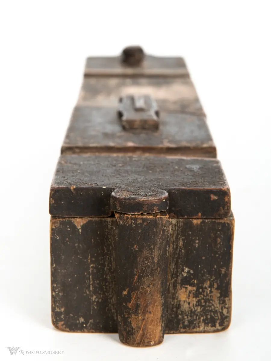 Rektangulær stokkeske hulet ut av et stykke tre. Dreielokk som har en sperreplate som dreier om en tapp og låses mot en låsetapp som holder hovedlokket låst mot en tapp på motsatt side.
