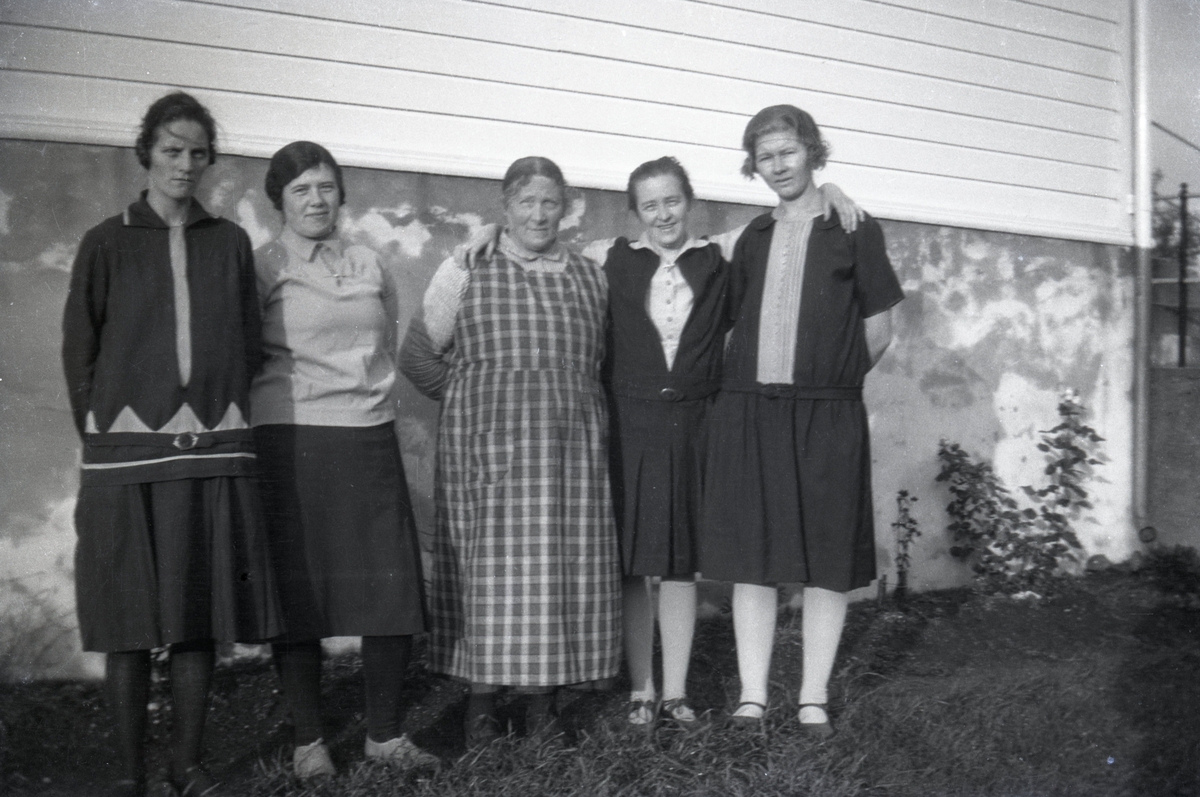 Gruppeportrett av fem kvinner foran en husvegg.