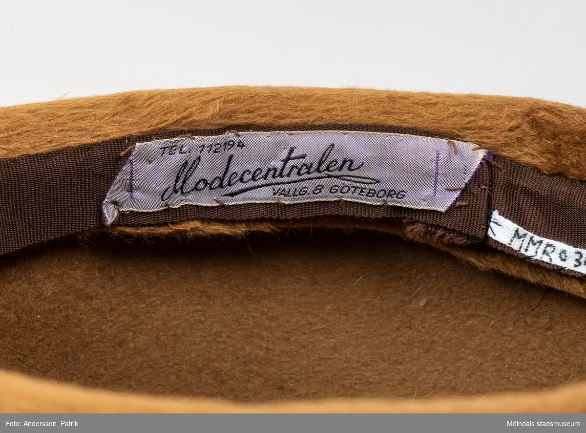 Ljusbrun hatt i ett pälsliknande tyg, från Modecentalen på Vallgatan i Göteborg. Hattens innerfoder är av filt. På insidan finns ett sidenband runtom och en fastsydd liten kam för att hålla hatten på plats. Där finns även en fastsydd vit etikett med texten:
"TEL. 11 21 94
Modecentralen
VALLG 8 GÖTEBORG"