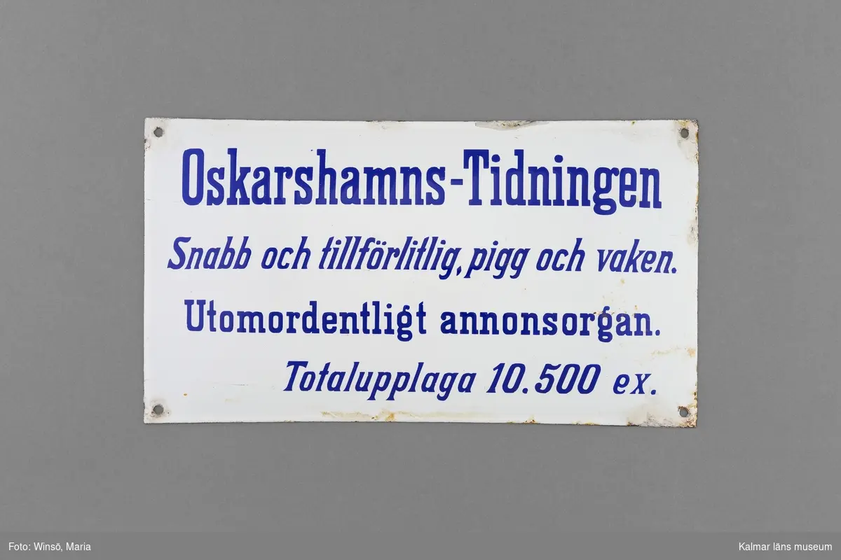 KLM 46449. Skylt, av järn. Vit emaljerad skylt med blå text som följer: Oskarshamns-Tidningen Snabb och tillförlitlig, pigg och vaken. Utomordentligt annonsorgan. Totalupplaga 10500 ex.