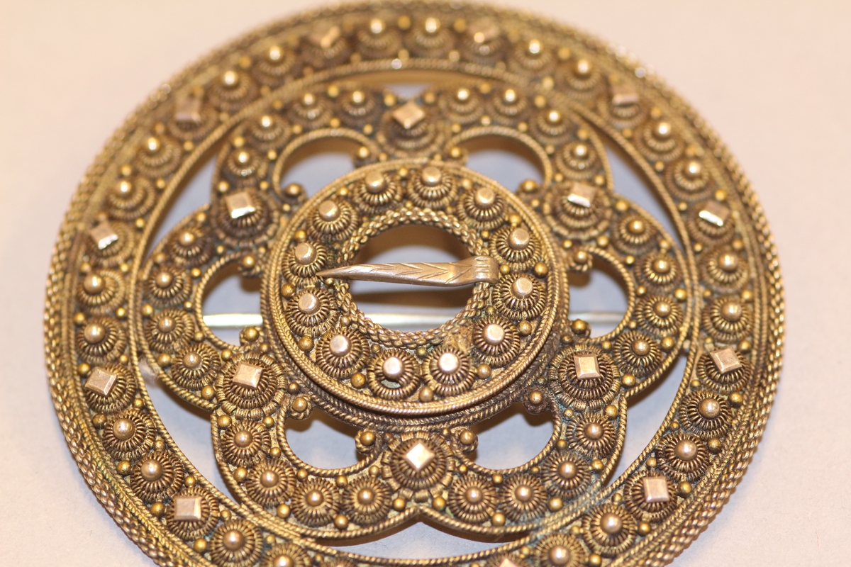 Ein kruse med plate, to krusar med knupp kring ytterste ringen. Inni slangen er det delingskrusar med plater oppå, fire kruser med knupp mellom desse. Rundt spretteholet ei rad med krusar og rund, litt større og flatare knupp. Spretteholet med torn. Sylgja er litt kuva.Nål på baksida.