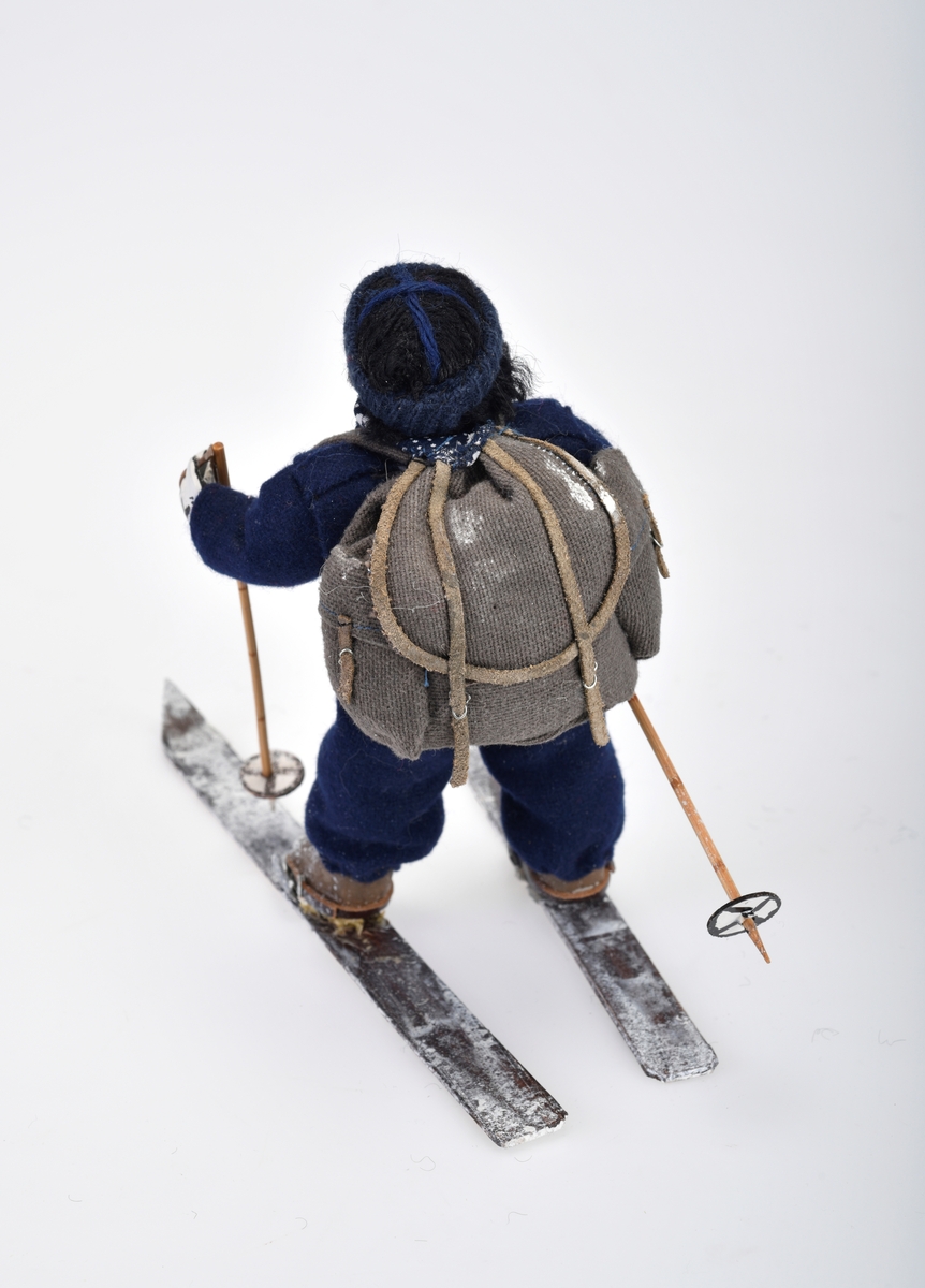 Liten damefigur med ryggsekk, ski på beina og skistaver i hendene.