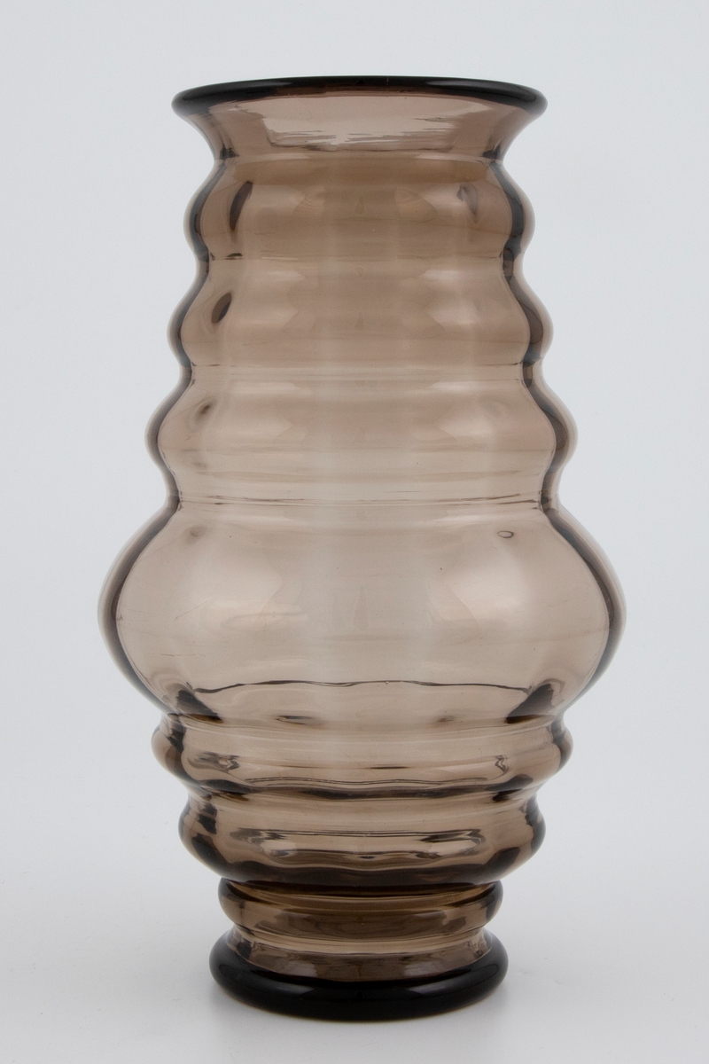 Balusterformet vase i bruntonet gjennomskinnelig glass med optisk dekor. Korpus er bygget opp av vulster som er bredest på midten av vasen, og smalner gradvis mot fot og munningsrand. Puntemerke under bunnen.