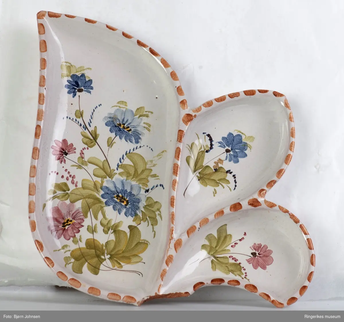 Keramikkfat formet som et blad, med ett stort og to små rom. Dekorert med håndmalte blomster. Stilken på bladet er knekt av og mangler. Antakelig produsert av Arol keramikk i Halden.