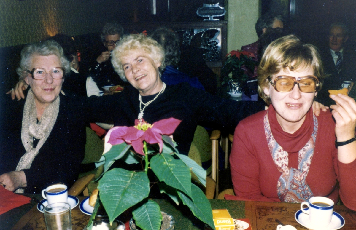 Tre kvinnor sitter till bords och dricker kaffe i Brattåsgårdens hobbylokal på Vommedalsvägen, okänt årtal. På bordet finns röd duk och röd julblomma.