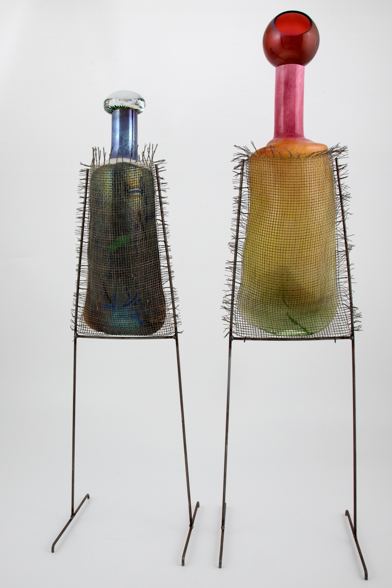 To skulpturer i glass i ulik høyde. Flaskeformet korpus i opakt glass i ulike farger. Sylindrisk hals med tilhørende glasspropp. Begge flaskene er innkapslet i et nettingshylster og bæres av et tobent stativ i metall.