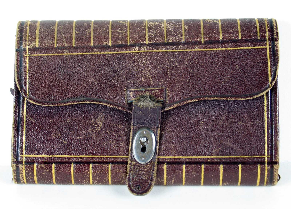 Lommebok med regnskapsbok i mørkt lilla skinn dekorert med gullstriper. Det er et åpent dobbelt rom og et lukket. Holder til blyant og notisblokk hvor Karoline førte regnskap.