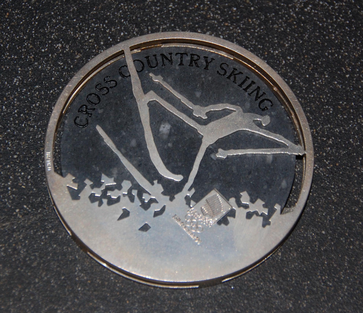 Medalje av metall og stein. På den ene siden er det motiv av de olympiske ringene over krystallmønster. På den andre siden er det piktogram av en skiløper og emblemet for de olympiske vinterleker på Lillehammer i 1994. Medaljen er plassert på et stativ av granitt.