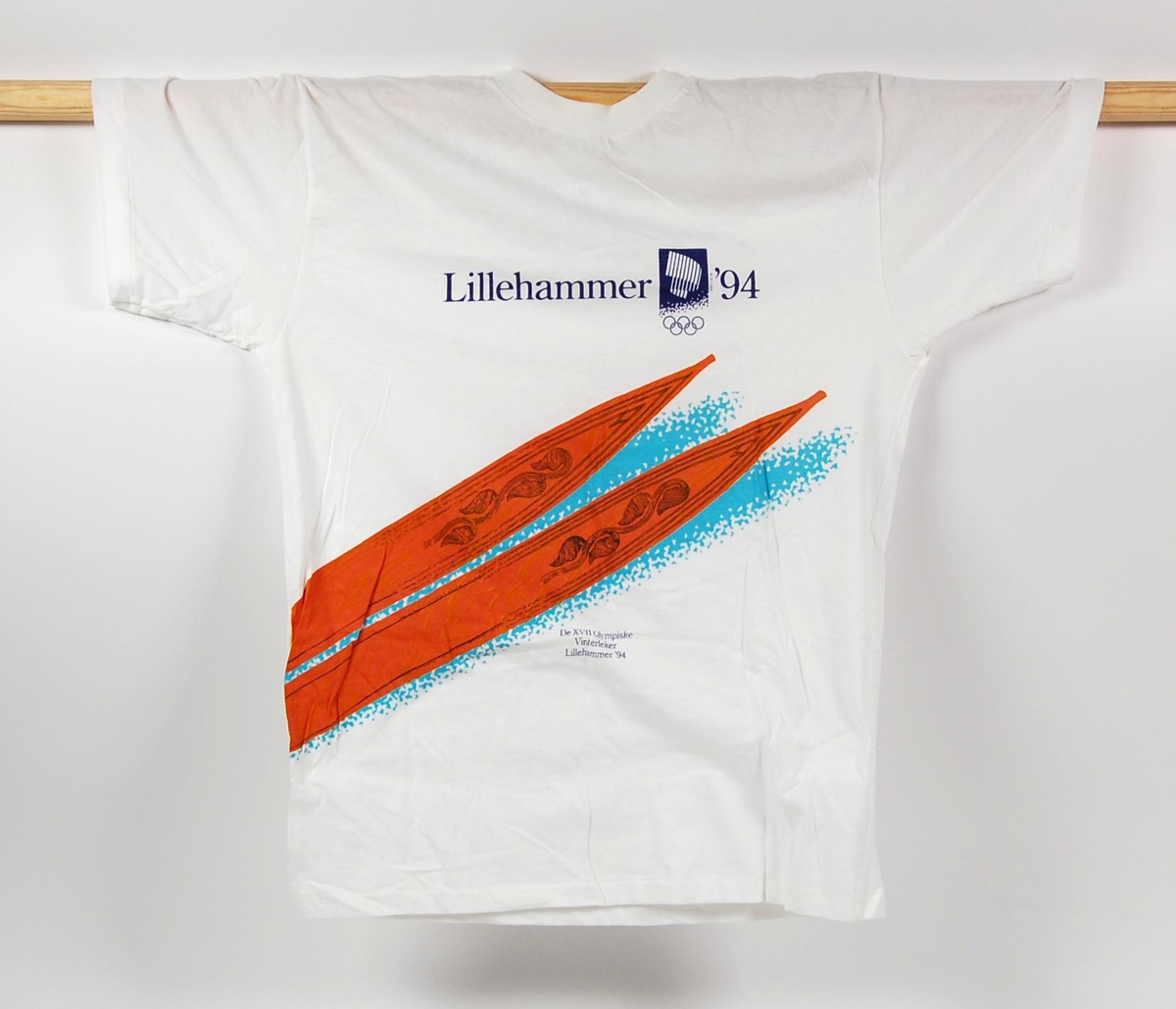 Hvit t-skjorte i størrelse L. Foran på t-skjorten er det en laminert logo for de olympiske vinterleker på Lillehammer i 1994 og et motiv av tuppene på et par treski av gammel modell.