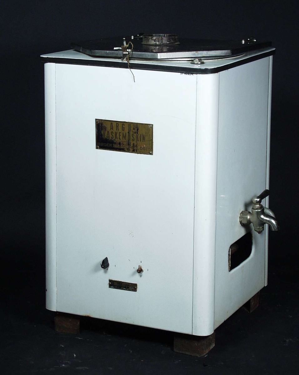 En hvit vaskemaskin med et sekskantet lokk i metall.
Den er uten vrimaskin og har to kraner og to brytere.