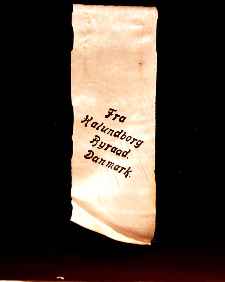 Bånd i kremfarget silke. På båndet står: Fra Kalundborg Byråd. Danmark. Båndet er kuttet i begge ender.
