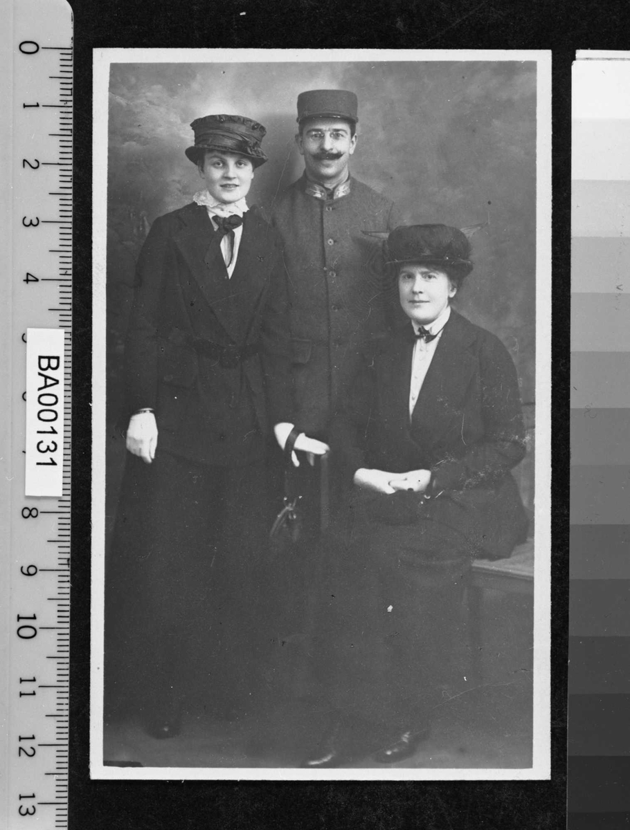 Fotografi av en mann i uniform og to damer, den yngste stående og den eldre sittende på benk i atelier.
Alle ser direkte på betrakteren.