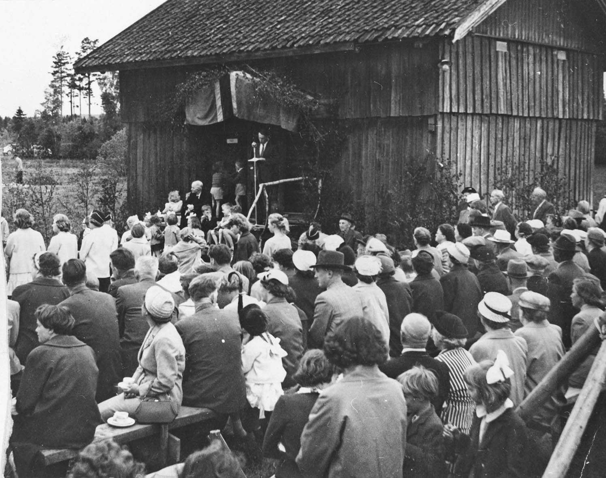 Historie-stevne på Nordby gård,folk er samlet på benker ute. Talerstol med norsk flagg og bjørkeløv langs stabbursveggen