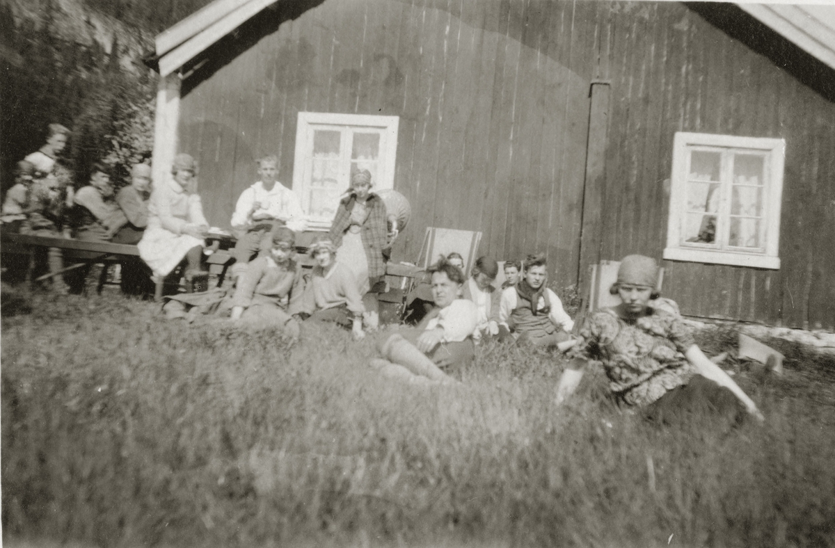 En gruppe mennesker sitter i solveggen og i gresset. Grammofon i bakgrunnen